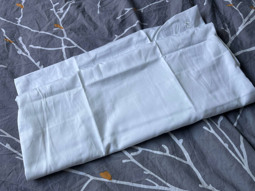 Fronha de algodão dobrada sobre si, num fundo de roupa de cama cinzenta escura com padrão